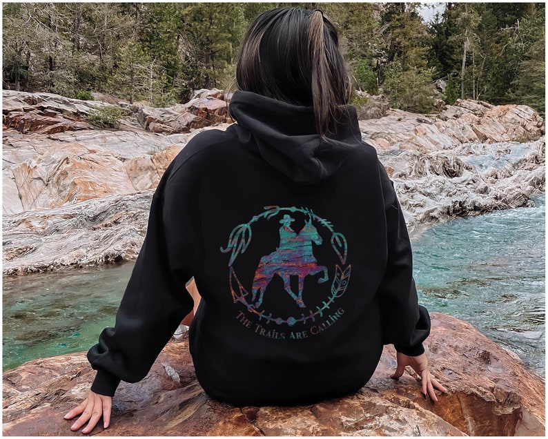 May include: Black hooded sweatshirt with a colorful graphic of a person riding a horse. The graphic is surrounded by a circle of leaves and the text "The Trails Are Calling".