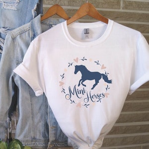 Może przedstawiać: Biała koszulka z niebieską sylwetką konia w wianku z r&oacute;żowych serc i liści. Pod koniem napis "Mini Horses".