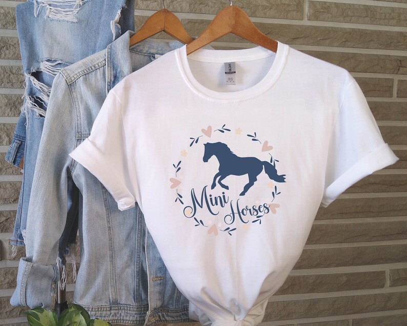 Może przedstawiać: Biała koszulka z niebieską sylwetką konia w wianku z r&oacute;żowych serc i liści. Pod koniem napis "Mini Horses".