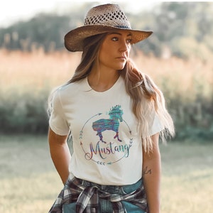 May include: White t-shirt with a colorful graphic of a horse and the word "Mustang" in a circle with arrows. The person wearing the shirt is wearing a brown cowboy hat and a plaid shirt tied around their waist.