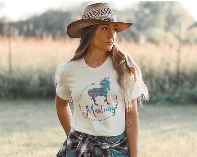 May include: White t-shirt with a colorful graphic of a horse and the word "Mustang" in a circle with arrows. The person wearing the shirt is wearing a brown cowboy hat and a plaid shirt tied around their waist.