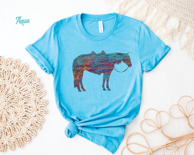 Op de afbeelding: Een lichtblauw t-shirt met een kleurrijk silhouet van een paard dat een zadel draagt. Het woord "Aqua" staat in de linkerbovenhoek van de afbeelding.