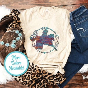May include: A natural-coloured t-shirt featuring a graphic of a person riding a horse inside a circular lasso frame with feathers. The graphic has a multi-coloured, textured pattern. A turquoise stone necklace, brown leather boots, and dark blue jeans are included.