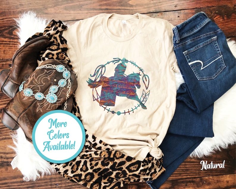 May include: A natural-coloured t-shirt featuring a graphic of a person riding a horse inside a circular lasso frame with feathers. The graphic has a multi-coloured, textured pattern. A turquoise stone necklace, brown leather boots, and dark blue jeans are included.