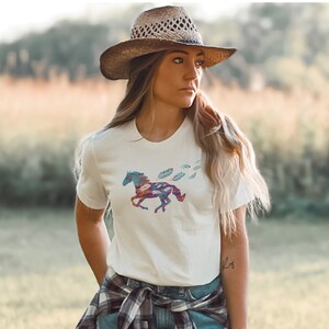 Peut inclure: Un t-shirt blanc avec un motif de cheval color&eacute; et des plumes. Le cheval est compos&eacute; de diff&eacute;rents motifs et couleurs, notamment du bleu, du rouge et du vert. La femme porte un chapeau de paille brun et une chemise &agrave; carreaux nou&eacute;e autour de sa taille.
