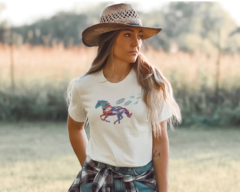 Peut inclure: Un t-shirt blanc avec un motif de cheval color&eacute; et des plumes. Le cheval est compos&eacute; de diff&eacute;rents motifs et couleurs, notamment du bleu, du rouge et du vert. La femme porte un chapeau de paille brun et une chemise &agrave; carreaux nou&eacute;e autour de sa taille.
