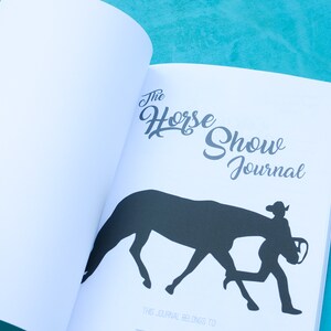 May include: The Horse Show Journal, a white journal with a black silhouette of a horse and rider on the cover. The text "The Horse Show Journal" is written in a cursive font.