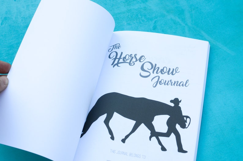 May include: The Horse Show Journal, a white journal with a black silhouette of a horse and rider on the cover. The text "The Horse Show Journal" is written in a cursive font.