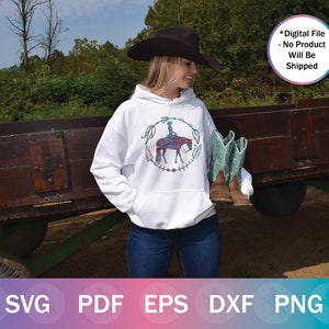 May include: White hoodie with a colorful graphic of a person riding a horse. The graphic is surrounded by a wreath of feathers and beads. The hoodie is being worn by a person wearing a black cowboy hat and blue jeans.
