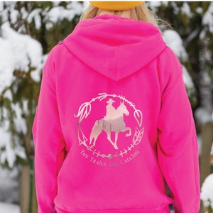 May include: A pink hoodie with a silver graphic of a person riding a horse with the text "The Trails Are Calling" on the back.
