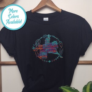 May include: A black short-sleeved t-shirt featuring a central graphic of a person riding a horse, enclosed within a circular frame of feathers and arrows. The graphic displays a textured, multi-coloured pattern of teal, orange, and purple hues.