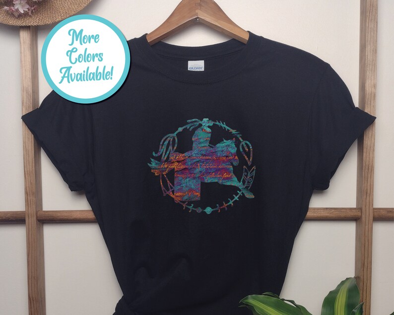 May include: A black short-sleeved t-shirt featuring a central graphic of a person riding a horse, enclosed within a circular frame of feathers and arrows. The graphic displays a textured, multi-coloured pattern of teal, orange, and purple hues.