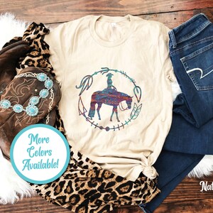 Pode incluir: Uma camiseta bege com um gr&aacute;fico de uma mulher montando um cavalo em um c&iacute;rculo com penas e outros desenhos. O texto "Mais cores dispon&iacute;veis!" est&aacute; em um c&iacute;rculo azul.