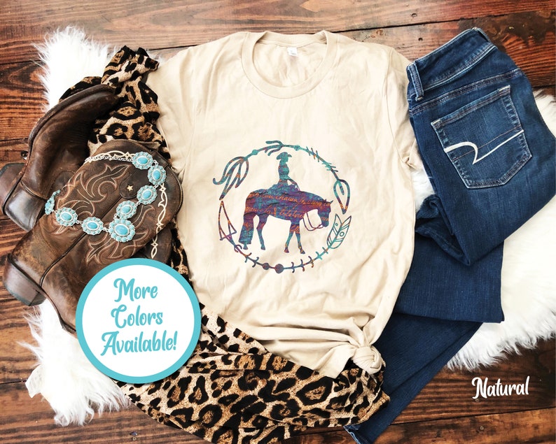 Pode incluir: Uma camiseta bege com um gr&aacute;fico de uma mulher montando um cavalo em um c&iacute;rculo com penas e outros desenhos. O texto "Mais cores dispon&iacute;veis!" est&aacute; em um c&iacute;rculo azul.