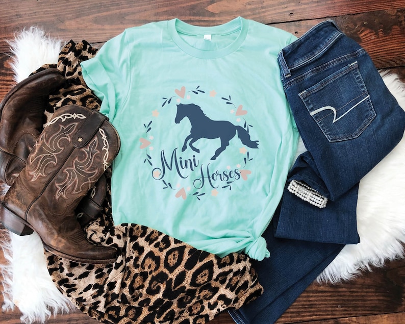 Może przedstawiać: Turkusowy t-shirt z grafiką ciemnoniebieskiego konia w wianku kwiatowym z napisem "Mini Horses". Koszulka leży na białym, puszystym podłożu z brązowymi kowbojskimi butami i niebieskimi dżinsami.