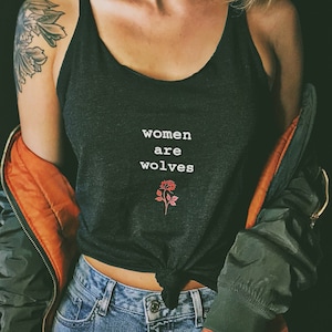 May include: A woman wearing a black tank top with the text "women are wolves" and a small rose graphic. She is also wearing a green jacket and blue jeans.