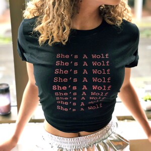 May include: A black t-shirt with the text "She's A Wolf" repeated multiple times in pink lettering.