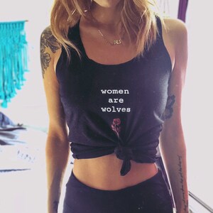 May include: A woman wearing a black tank top with the text "women are wolves" and a small rose graphic. She is also wearing black leggings.