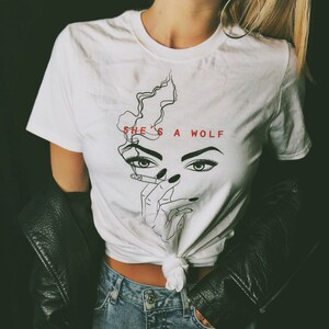 Op de afbeelding: Wit T-shirt met een zwart-witte lijntekening van een vrouwenhoofd met een sigaret in haar hand. De tekst "SHE'S A WOLF" is in rood boven de tekening gedrukt.