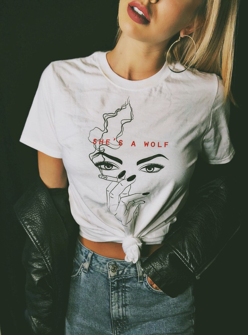 Op de afbeelding: Wit T-shirt met een zwart-witte lijntekening van een vrouwenhoofd met een sigaret in haar hand. De tekst "SHE'S A WOLF" is in rood boven de tekening gedrukt.