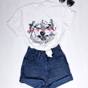 May include: White t-shirt with a black and white wolf graphic and the text "she's a wolf" in red. The t-shirt is paired with blue denim shorts.