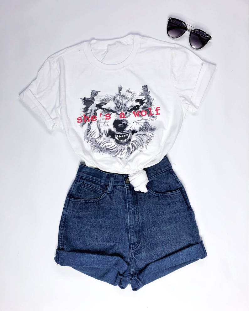 May include: White t-shirt with a black and white wolf graphic and the text "she's a wolf" in red. The t-shirt is paired with blue denim shorts.