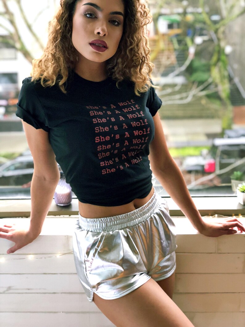 May include: A woman wearing a black t-shirt with the text "She's A Wolf" repeated multiple times and silver metallic shorts.