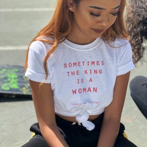 May include: A white t-shirt with the text "Sometimes the king is a woman" in red lettering. The shirt is tied at the waist and the model is wearing black pants with a yellow stripe.