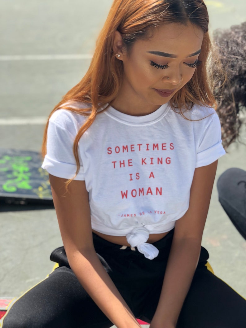May include: A white t-shirt with the text "Sometimes the king is a woman" in red lettering. The shirt is tied at the waist and the model is wearing black pants with a yellow stripe.