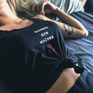 May include: A black tank top with the text "women are wolves" printed on the front. A red rose is printed below the text. The tank top is tied at the front.