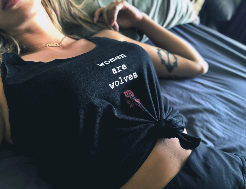 May include: A black tank top with the text "women are wolves" printed on the front. A red rose is printed below the text. The tank top is tied at the front.