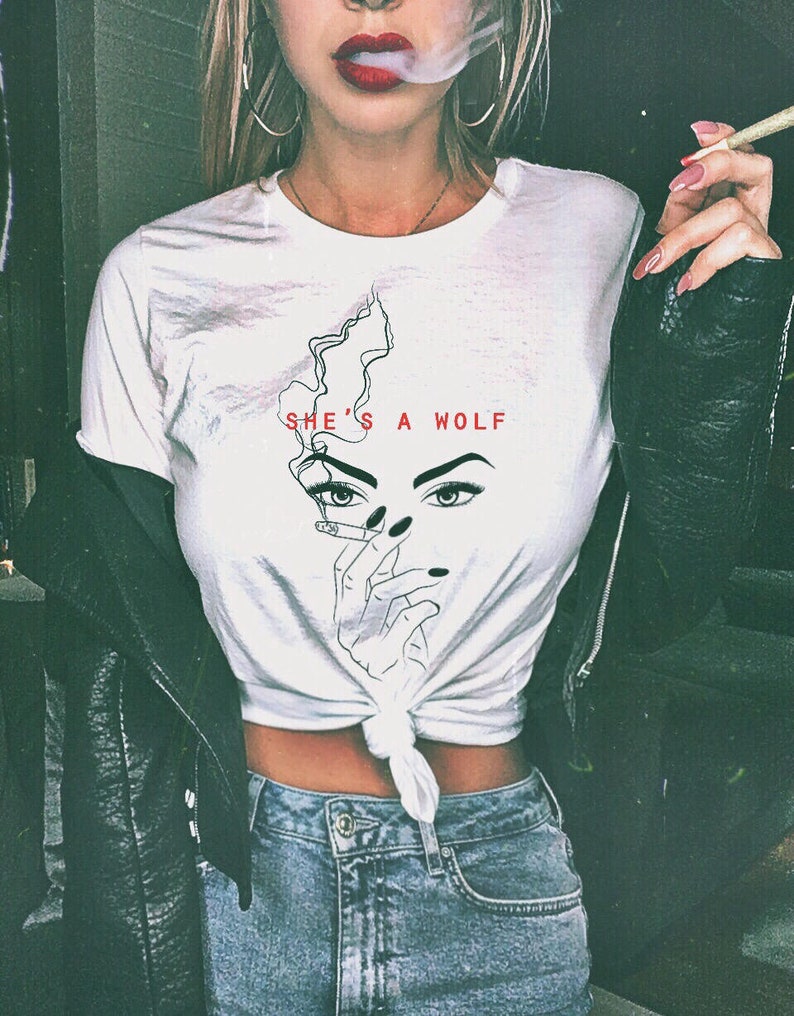 Op de afbeelding: Een wit T-shirt met een afbeelding van een vrouwenhoofd met een sigaret in haar hand. De tekst "SHE'S A WOLF" staat op het shirt gedrukt.
