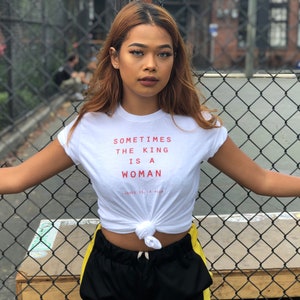 May include: A woman wearing a white t-shirt with the text "Sometimes the king is a woman" in red. She is standing in front of a chain-link fence and has her arms outstretched.