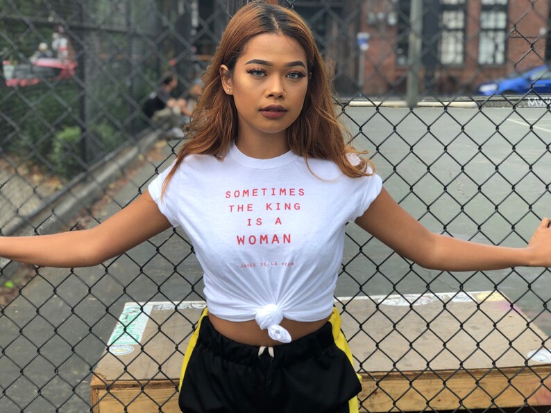 May include: A woman wearing a white t-shirt with the text "Sometimes the king is a woman" in red. She is standing in front of a chain-link fence and has her arms outstretched.