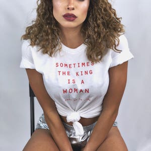 May include: A woman with curly brown hair is wearing a white t-shirt with the text "Sometimes the King is a Woman" in red. She is also wearing silver shorts.