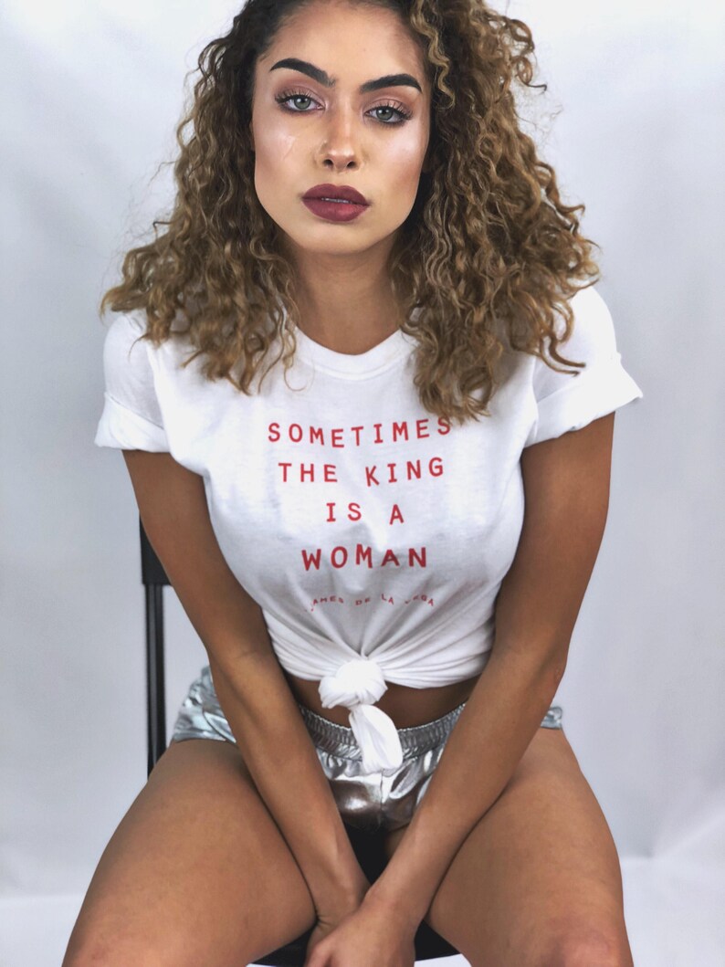 May include: A woman with curly brown hair is wearing a white t-shirt with the text "Sometimes the King is a Woman" in red. She is also wearing silver shorts.