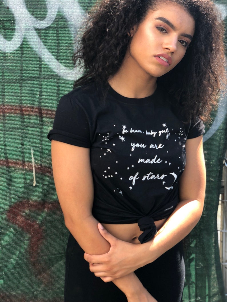 May include: A black t-shirt with a white graphic that says "be brave, baby girl you are made of stars" with a crescent moon and stars around the text.