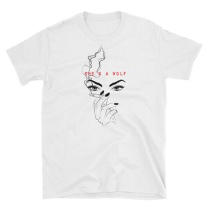 Op de afbeelding: Wit T-shirt met een zwart-witte lijntekening van een vrouwen gezicht met een sigaret en de tekst "SHE'S A WOLF" in rood.