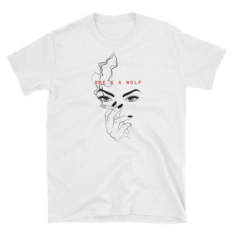 Op de afbeelding: Wit T-shirt met een zwart-witte lijntekening van een vrouwen gezicht met een sigaret en de tekst "SHE'S A WOLF" in rood.