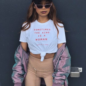 May include: A woman wearing a white t-shirt with the text "Sometimes the king is a woman - James de la Vega" in red. She is also wearing a black baseball cap, pink sunglasses, a purple bomber jacket, and brown shorts with a gold chain belt.