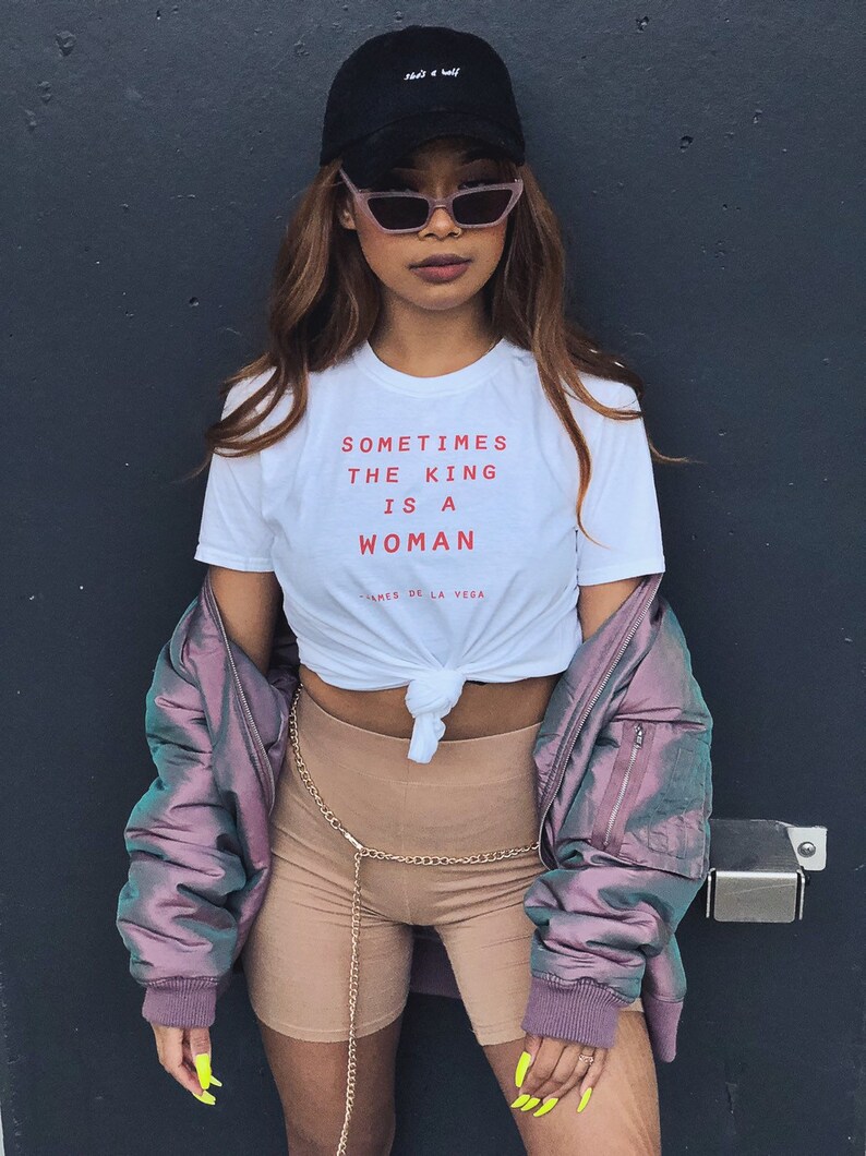 May include: A woman wearing a white t-shirt with the text "Sometimes the king is a woman - James de la Vega" in red. She is also wearing a black baseball cap, pink sunglasses, a purple bomber jacket, and brown shorts with a gold chain belt.
