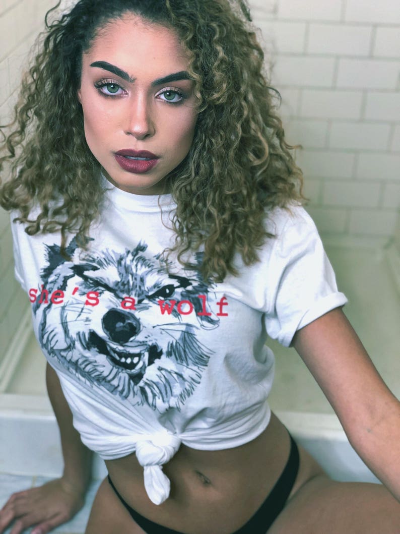 May include: A white t-shirt with a wolf graphic and the text "she's a wolf" in red. The shirt is tied at the waist.