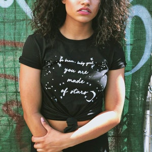 May include: A black t-shirt with a white graphic that says "be brave, baby girl, you are made of stars". The shirt is tied at the waist and the model is wearing black trousers.