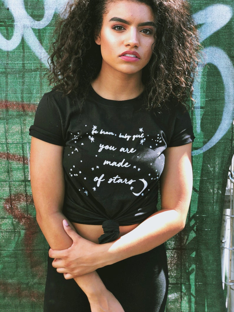 May include: A black t-shirt with a white graphic that says "be brave, baby girl, you are made of stars". The shirt is tied at the waist and the model is wearing black trousers.