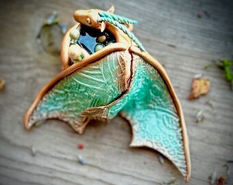 Könnte beinhalten: Eine kleine, grüne und goldene Drachenskulptur mit ausgebreiteten Flügeln. Der Drache ist in einem Nest aus Perlen zusammengerollt und hat eine detaillierte, strukturierte Oberfläche.