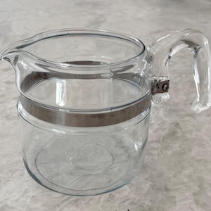 Puede incluir: Una jarra pequeña o lechera de vidrio transparente con un cuerpo liso y un pico vertedor ancho. Una banda metálica de tono plateado rodea la parte superior del recipiente, y cuenta con un asa resistente integrada de vidrio transparente.