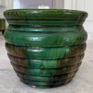 May include: A green ceramic pot with a ribbed design. The pot has a glossy finish and a rounded shape. The color is a deep, rich green with hints of brown. The pot is likely used for decorative purposes or for holding plants.