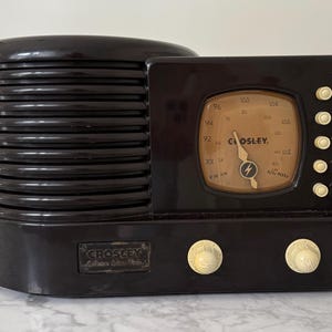 May include: A dark brown Crosley radio featuring a horizontal ribbed speaker grille on the left and a square tuning dial with a tan background on the right. The device includes five small cream-coloured buttons, two round control knobs, and a Crosley logo.