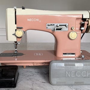 May include: A vintage Necchi Lelia 514 sewing machine in a two-tone dusty pink and cream finish. The machine features a metal body, a stitch length adjustment lever, a circular dial, and a black foot pedal. A grey plastic Necchi case is included.