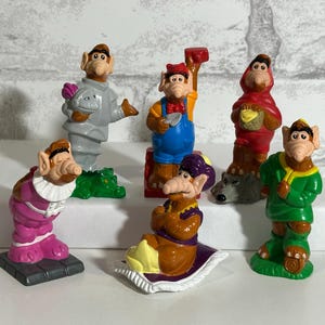 May include: A collection of colorful figurines of a cartoon character in various outfits. The figurines are in different poses and outfits, including a chef, a knight, and a character in a red hooded cloak. The figurines are on a white surface.